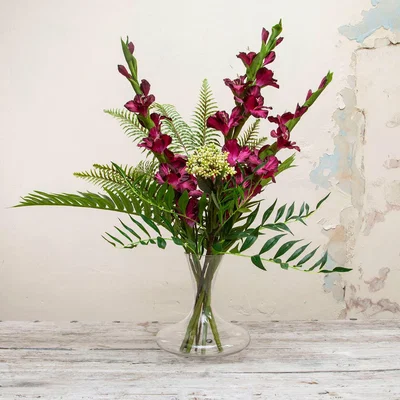 The Range Gladioli with Ferns and Berries in a Decanter Glass Vase by Peony - Burgundy red with strong green leaves